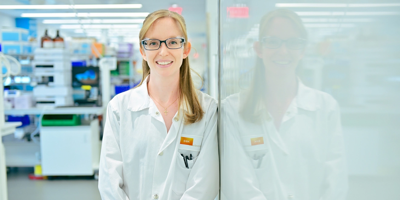 Nicole is smiling at the camera in a lab, wearing a lab coat