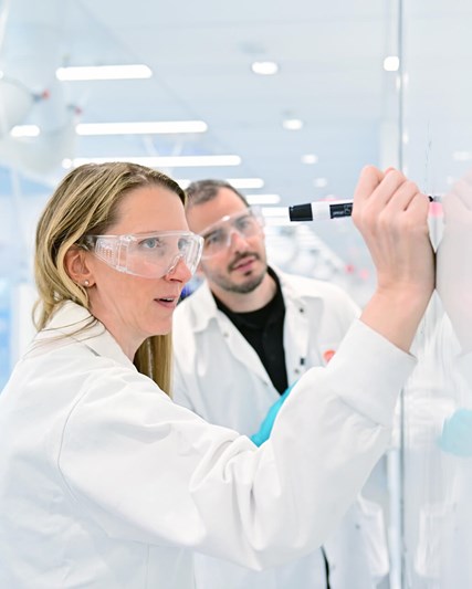 Two scientists in white coats and goggles collaborating while writing on a white board
