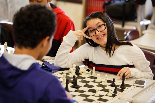 A teen boy and a girl playing chess. Young girl is smiling at the camera. 