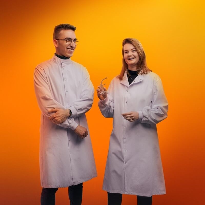 A man and a woman in lab coats laughing in front of an orange background