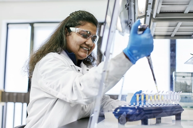 Scientist wearing goggles, white jacket, blue gloves using a pipette