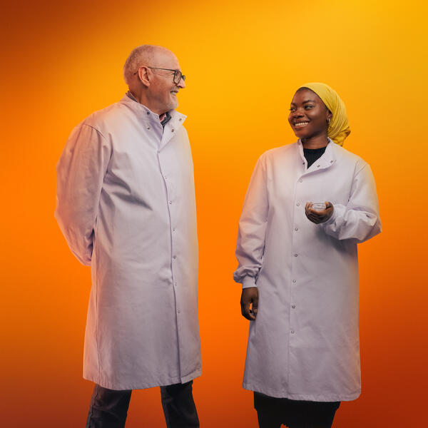 Two people in lab PPE smiling against an orange background