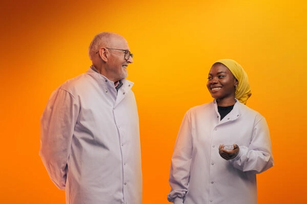 Two people in lab PPE smiling against an orange background