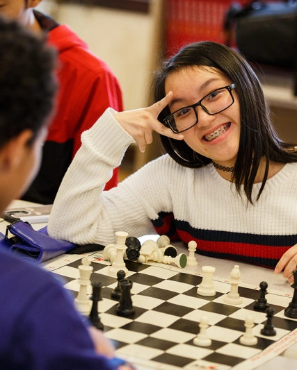 A teen boy and a girl playing chess. Young girl is smiling at the camera.